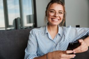 Woman in glasses and striped shirt smiling while holding her smartphone